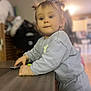 toddler, child, indoor, table, pigtails, curious, young_child, clothing, face, person, hands, wooden_table, soft_lighting, room, background_blur, expression, standing, casual, home, playful