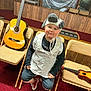apron, boy, carpet, casual_clothing, chair, child, costume, guitar, hat, indoors, musical_instrument, playful, portrait, red_carpet, room, shorts, smiling, sneakers, ukulele, wood_paneling