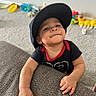 toddler, child, cap, black_tshirt, red_trim, carpet, toys, colorful, wall, electrical_outlet, hand, face, smile, indoor, furniture, playroom, baby, person, expression, cheeky