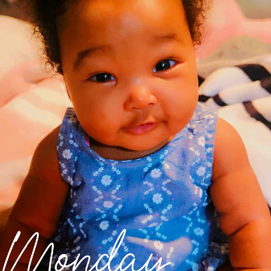 Richael joined the competition — help win amazing prizes! baby, child, curly_hair, blue_dress, floral_pattern, bed, blanket, headboard, smile, portrait, indoors, cute, infant, person, sitting, skin, hand, face, eyes, young