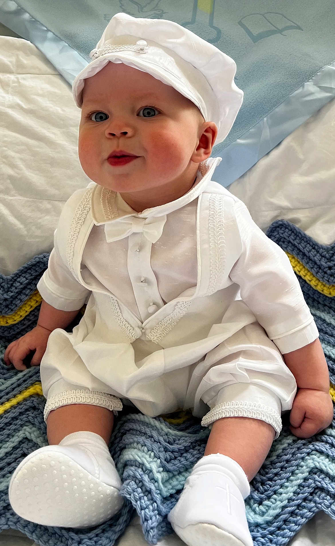Christopher is registered to the contest to win money with this photo: baby, child, white_clothing, hat, sitting, blanket, knitted_blanket, indoors, cute, portrait, smiling, footwear, socks, face, blue_eyes, infant, hands, cheeks, cute_expression, soft_lighting