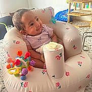 Reyna is registered to the contest to win money with this photo: baby, infant, inflatable_seat, toy, colorful_toy, indoor, child, happy, smiling, seated, patterned_floor, wooden_shelf, plant_pot, purple_clothing, face, cute, small, home, decor, playful