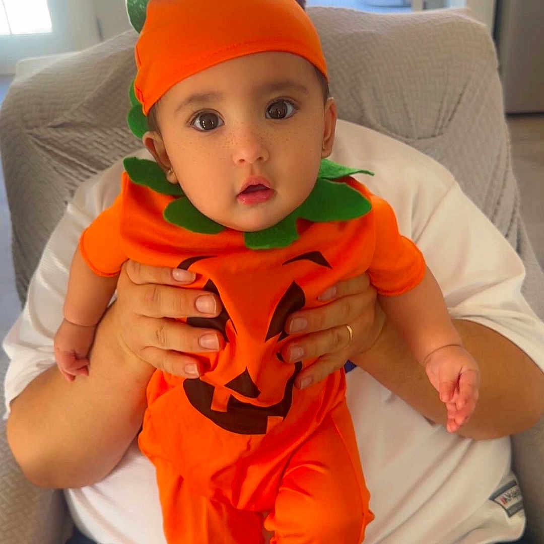 Reyna joined the competition — help win amazing prizes! baby, child, infant, halloween, pumpkin_costume, orange, green, jack_o_lantern, hat, costume, sitting, held, portrait, wide_eyed, indoors, chair, sunlight, doorway, family, cute