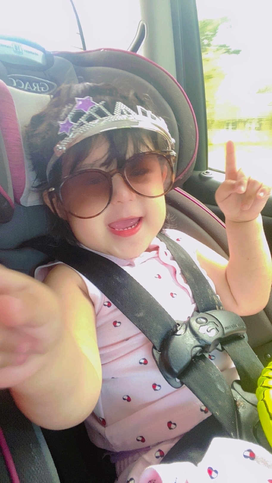 Neisly joined the competition — help win amazing prizes! toddler, child, car_seat, sunglasses, tiara, smile, happy, pointing, seatbelt, clothing, car_interior, window, daylight, cute, fun, baby, face, person, sitting, accessory