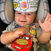 Jaxson is registered to the contest to win money with this photo: baby, child, smiling, car_seat, tshirt, burger_king_crown, hand, toy, bottle, happy, cute, indoor, infant, clothing, seatbelt, portrait, face, expression, colorful, playful