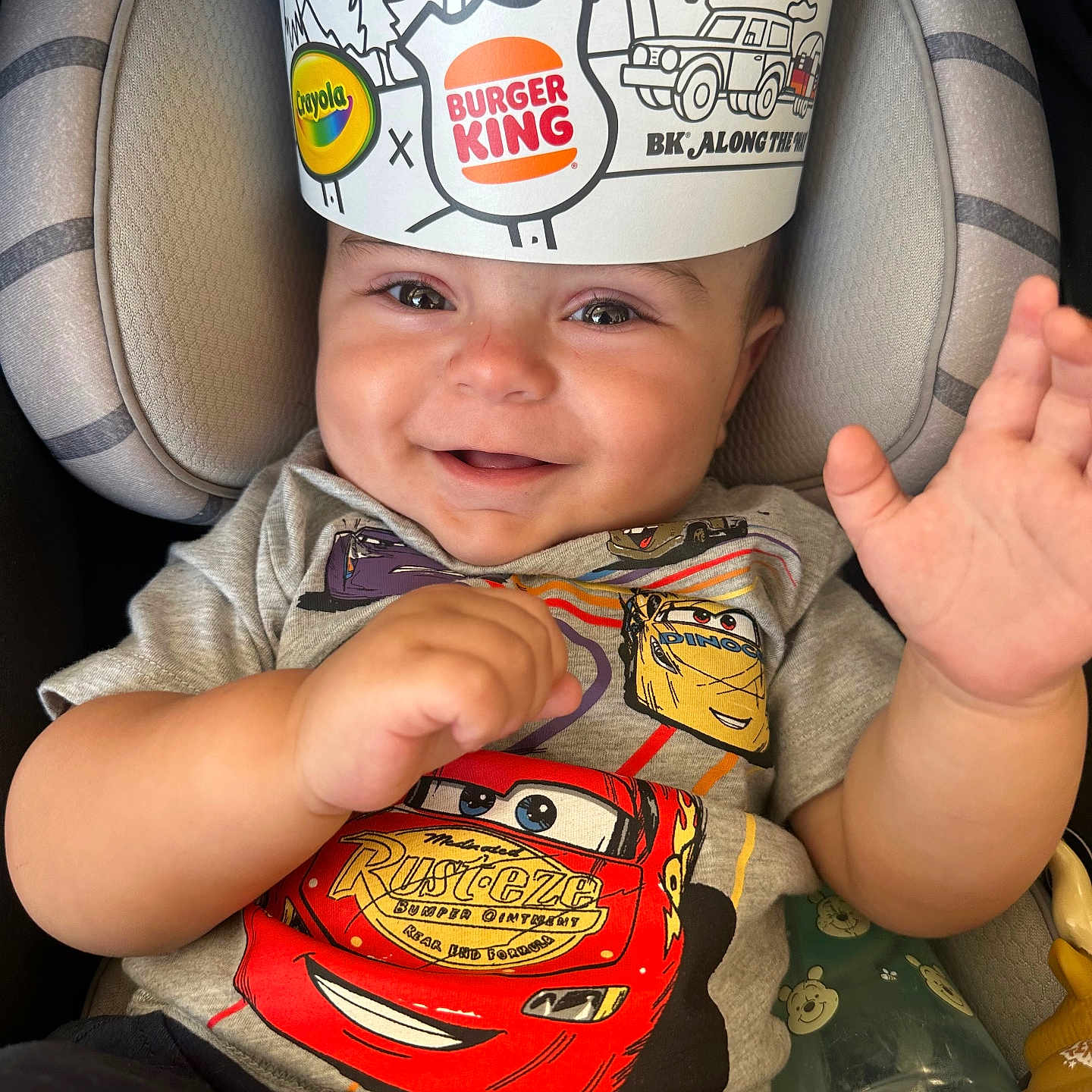 Jaxson is registered to the contest to win money with this photo: baby, child, smiling, car_seat, tshirt, burger_king_crown, hand, toy, bottle, happy, cute, indoor, infant, clothing, seatbelt, portrait, face, expression, colorful, playful