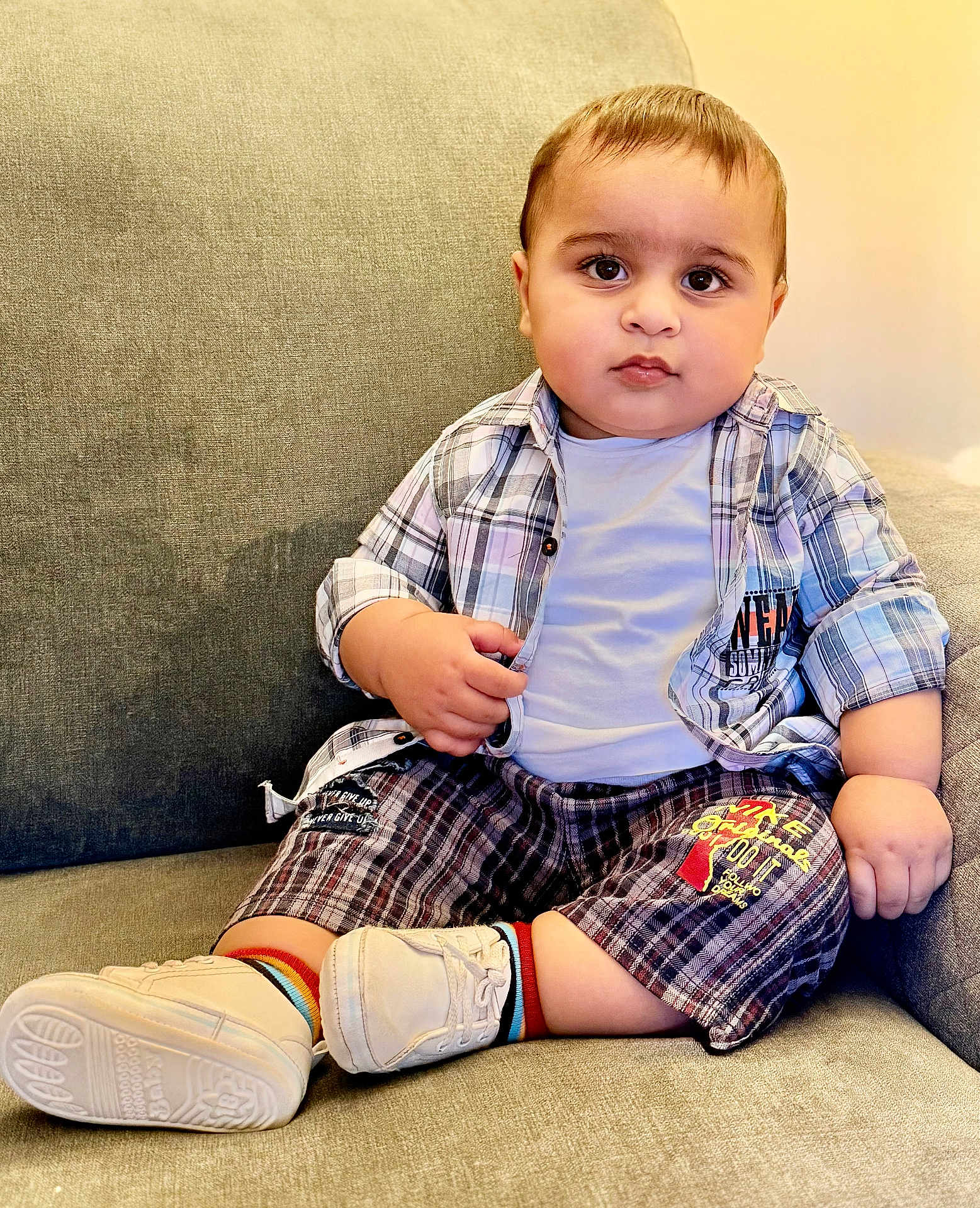 Muhammad Aahill is registered to the contest to win money with this photo: baby, toddler, child, plaid_shirt, white_tshirt, plaid_shorts, rainbow_socks, white_shoes, couch, indoor, seated, curious, chubby_cheeks, person, portrait, casual_clothing, comfortable, young_child, home, cute