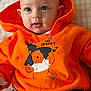 baby, child, hoodie, orange_clothing, winnie_the_pooh, halloween, bat, cushion, pillow, text, face, eyes, sitting, black_pants, white_stripes, indoor, cute, infant, portrait, smiling