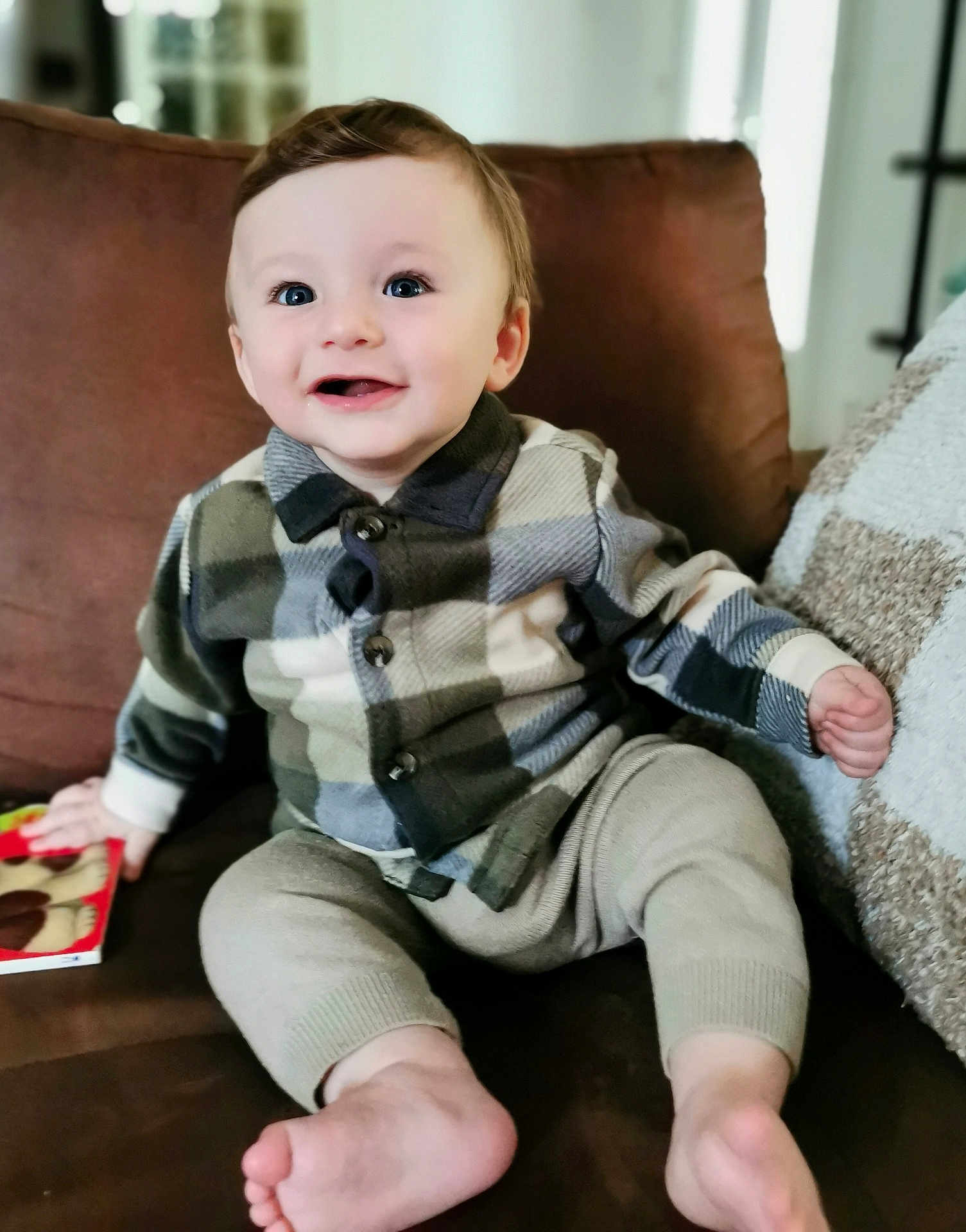 Aaron a rejoint le concours — aidez-le/la à gagner de superbes lots ! baby, child, smiling, plaid_shirt, beige_pants, barefoot, couch, indoor, happy, blue_eyes, holding_book, portrait, cute, seated, cozy, young_child, person, home, infant, comfortable