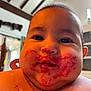 baby, face, food_stains, messy, indoor, child, curious, close_up, cheeks, bib, polka_dots, young_child, expression, head, person, portrait, warm_light, home_interior, dark_eyes, hair