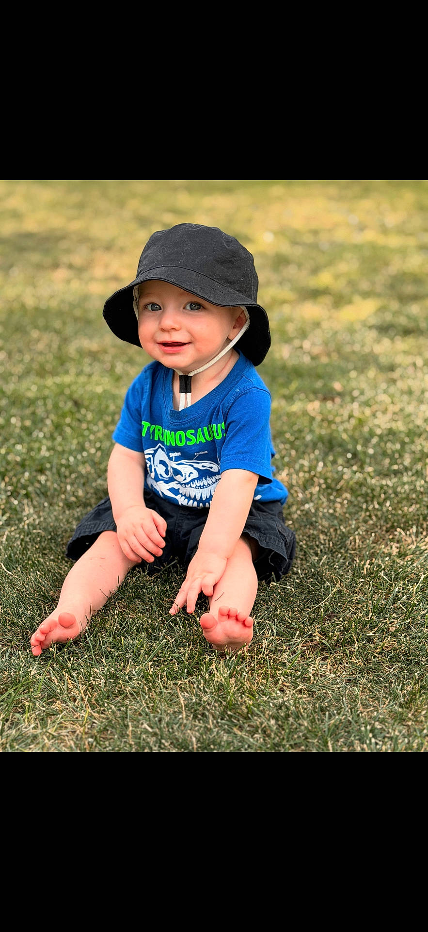 baby, barefoot, black_hat, blue_shirt, casual_clothing, child, cute, daylight, grass, green, happy, nature, outdoor, playful, portrait, sitting, smiling, sunny, toddler, young_child