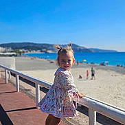 Alma a rejoint le concours — aidez-le/la à gagner de superbes lots ! beach, child, coast, daylight, dress, footwear, girl, ocean, outdoor, pigtails, playful, railing, sand, shadows, sky, smile, summer, sunny, toddler, vacation
