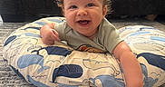 Felipe is registered to the contest to win money with this photo: baby, smiling, curly_hair, cushion, whale_pattern, pointing, living_room, carpet, sofa, blanket, happy, infant, child, indoor, cute, person, toddler, clothing, face, home