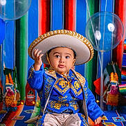 Martin is registered to the contest to win money with this photo: toddler, child, traditional_clothing, sombrero, blue_outfit, gold_embroidery, balloons, colorful_blanket, decorations, sitting, portrait, celebration, festive, cute, indoors, bright_colors, face, person, curious, hat