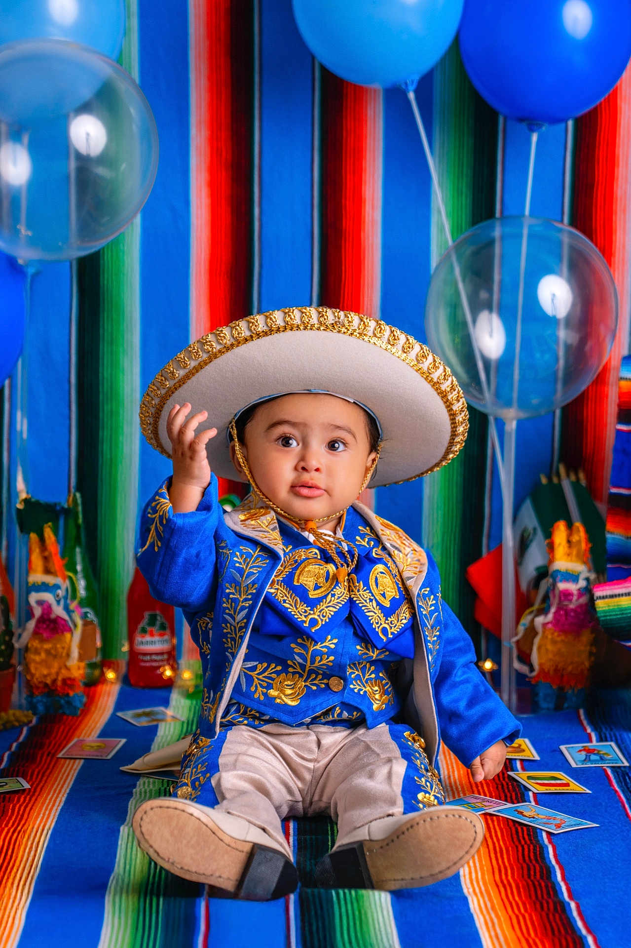 Martin is registered to the contest to win money with this photo: toddler, child, traditional_clothing, sombrero, blue_outfit, gold_embroidery, balloons, colorful_blanket, decorations, sitting, portrait, celebration, festive, cute, indoors, bright_colors, face, person, curious, hat