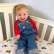 Kieran is registered to the contest to win money with this photo: baby, bare_feet, bedsheet, blond_hair, blue_eyes, child, construction_vehicles, crib, denim, furniture, happy, indoor, infant, nursery, overalls, photography, portrait, red_shirt, sitting, smiling