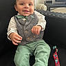 baby, child, smiling, sitting, clothing, vest, bow_tie, green_pants, white_shirt, couch, black_couch, toy, colorful_toy, face, happy, indoors, television, furniture, person, portrait