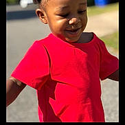 Amiri is registered to the contest to win money with this photo: child, toddler, person, portrait, smiling, happy, outdoor, sunlight, red_shirt, braided_hair, dark_skin, face, head, standing, walking, street, road, grass, blue_bin, phone_screenshot
