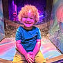child, toddler, curly_hair, smiling, play_structure, colorful, rainbow_lights, indoor, blue_shirt, shorts, happy, playground, reflection, glass, fun, cute, person, sitting, bright_colors, light_effects