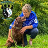 affection, blue_hoodie, casual_clothing, chocolate_labrador, collar, dog, glasses, grass, greenery, happy, kneeling, leash, logo, nature, outdoor, paw_print, person, pet, short_hair, smiling