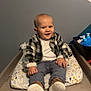Mathis a rejoint le concours — aidez-le/la à gagner de superbes lots ! baby, child, plaid_jacket, cushion, changing_table, shoes, indoor, smiling, cute, toddler, clothing, face, person, furniture, wooden_surface, toy, white_wall, blue_wall, patterned_cushion, happy