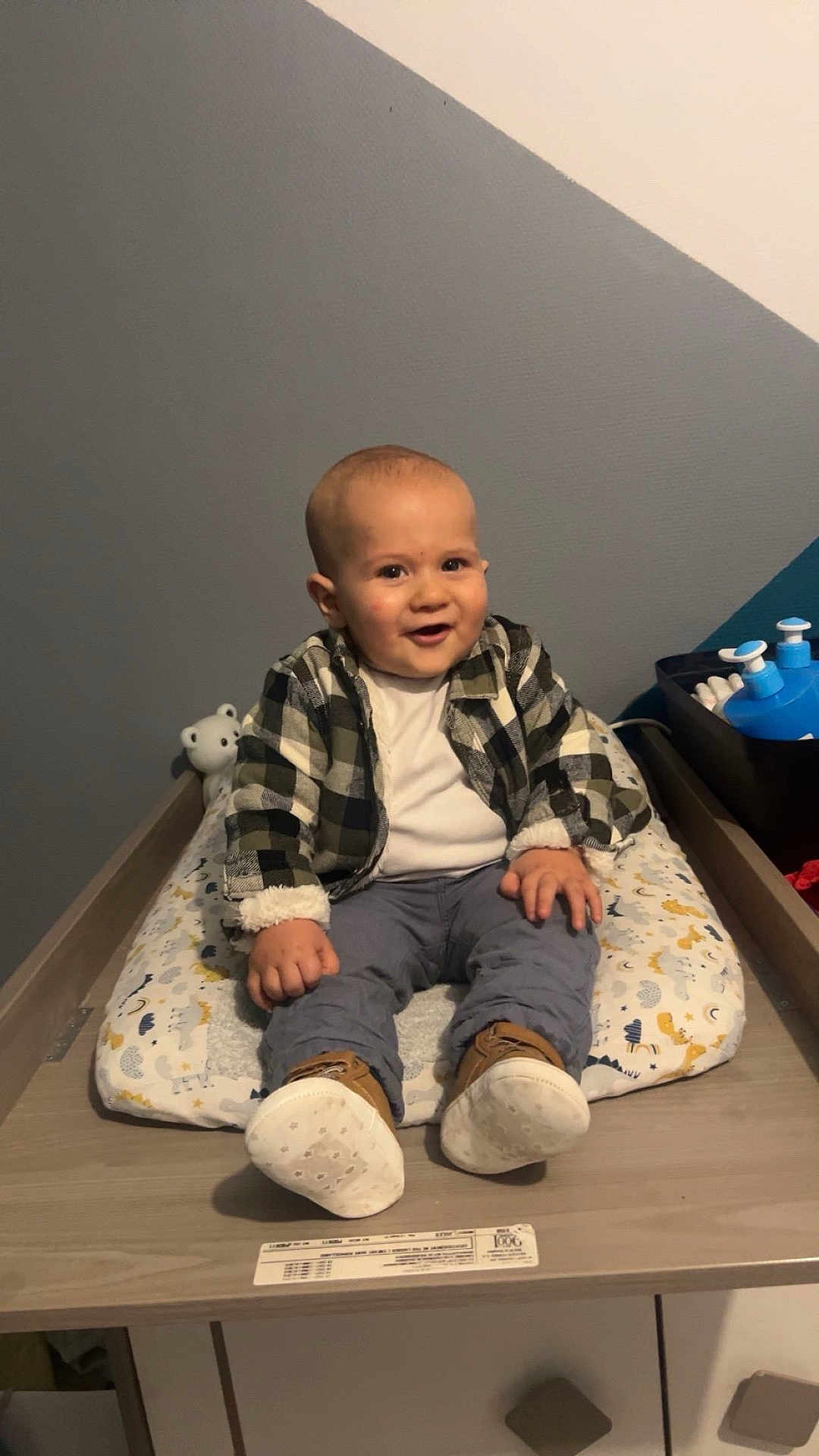 Mathis a rejoint le concours — aidez-le/la à gagner de superbes lots ! baby, child, plaid_jacket, cushion, changing_table, shoes, indoor, smiling, cute, toddler, clothing, face, person, furniture, wooden_surface, toy, white_wall, blue_wall, patterned_cushion, happy