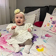 Madina a rejoint le concours — aidez-le/la à gagner de superbes lots ! baby, bed, blanket, child, comfort, cute, decor, floral_bedspread, flower, happy, headband, home, indoors, infant, person, pillow, sitting, smiling, soft_lighting, white_dress