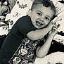 Colt is registered to the contest to win money with this photo: toddler, child, smiling, curly_hair, plush_toy, bed, dinosaur_sheets, black_and_white, happy, indoor, clothing, cartoon_characters, face, portrait, cute, lying_down, playful, young_child, carpet, cozy