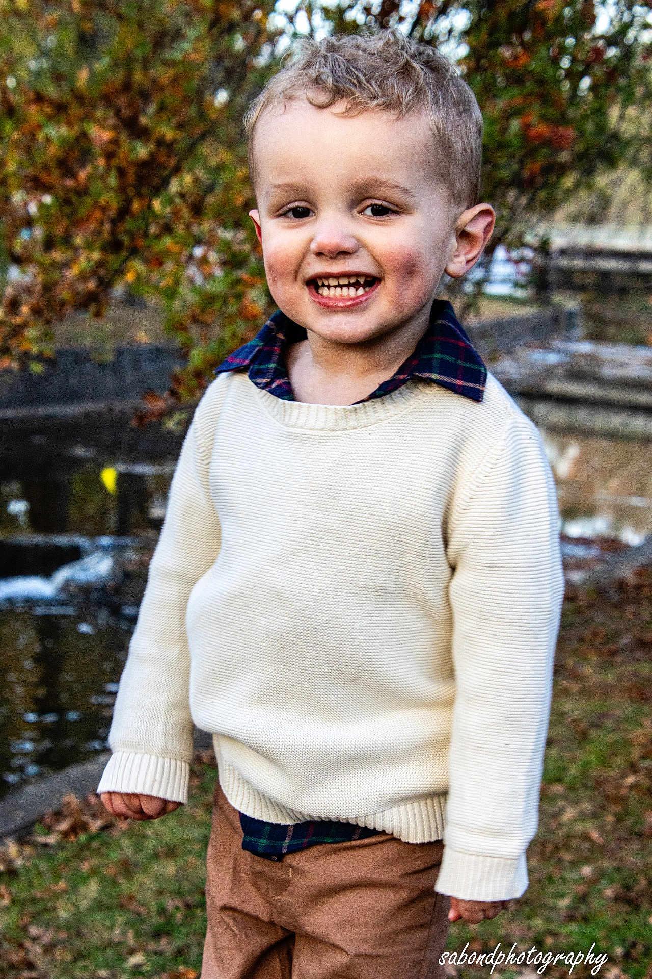 Colt is registered to the contest to win money with this photo: child, boy, smiling, outdoor, autumn, sweater, plaid_shirt, pants, grass, pond, water, trees, park, nature, person, fall_leaves, happy, young, curly_hair, portrait