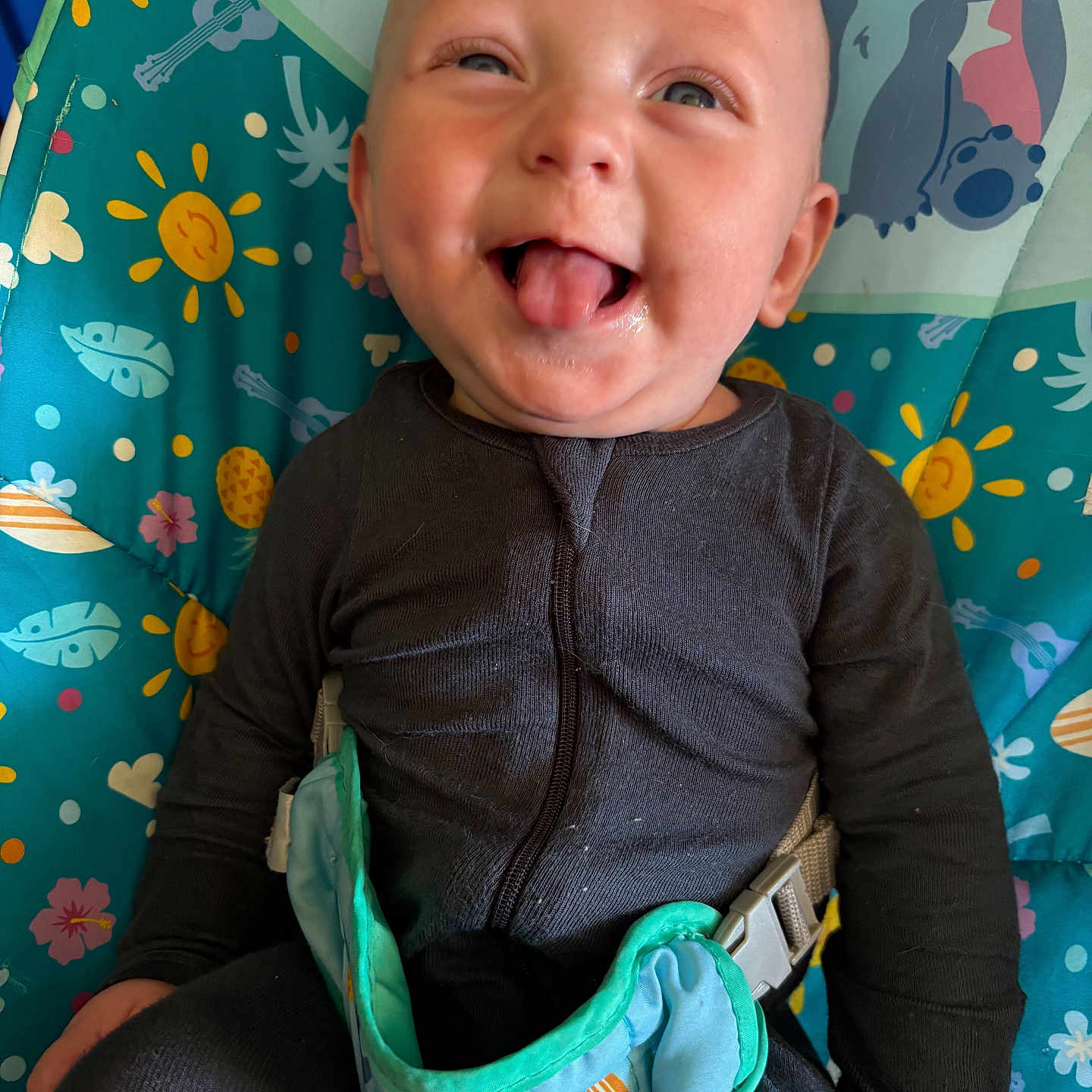 Oliver joined the competition — help win amazing prizes! baby, infant, child, smiling, tongue_out, sitting, portrait, face, eyes, black_clothing, jumpsuit, harness, seat, activity_seat, colorful, patterned_background, stitch, indoor, happy, playful