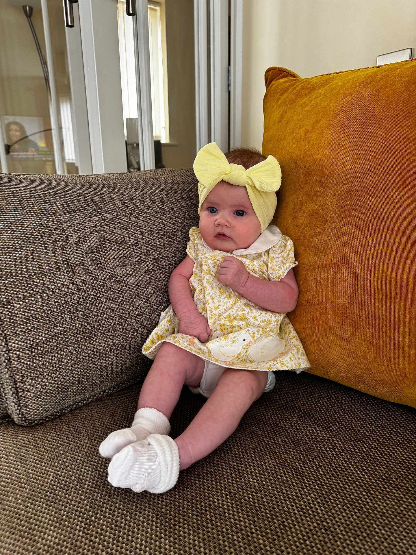 Averie-Bay joined the competition — help win amazing prizes! baby, child, infant, headband, yellow_bow, dress, socks, couch, cushion, living_room, furniture, indoors, portrait, seated, curious, soft_lighting, home, window, door, lamp