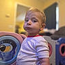 toddler, child, indoor, play_equipment, curious, puffed_cheeks, short_sleeve_shirt, blonde_hair, face, person, toy, colorful, blurred_background, home, expression, young_child, casual_clothing, portrait, standing, room