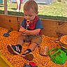 toddler, child, sandbox, corn_kernels, pacifier, toys, watering_can, measuring_cup, toy_vehicle, outdoor, grass, wooden_box, casual_clothing, shorts, sneakers, playtime, summer, daylight, person, background