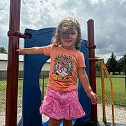 Annalise joined the competition — help win amazing prizes! child, playground, outdoors, smiling, portrait, play_structure, slide, sky, clouds, sunlight, pink_shorts, orange_shirt, cowboy_boots, curly_hair, standing, playground_equipment, fence, trees, backyard, summer