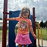 child, playground, outdoors, smiling, portrait, play_structure, slide, sky, clouds, sunlight, pink_shorts, orange_shirt, cowboy_boots, curly_hair, standing, playground_equipment, fence, trees, backyard, summer