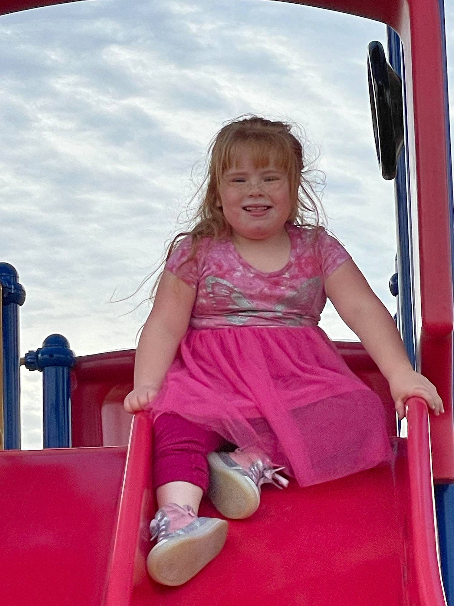 Willo is registered to the contest to win money with this photo: blond, child, chute, cloud, facial_expression, fun, happy, human_leg, joy, leisure, magenta, outdoor_play_equipment, person, pink, recreation, shorts, sitting, sky, smile, t_shirt