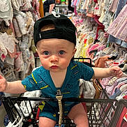 Holden is registered to the contest to win money with this photo: baby, shopping_cart, hat, clothing_store, baby_clothes, blue_onesie, person, child, retail, backwards_cap, shopping, cute, wide_eyes, indoor, rack, apparel, infant, store, toddler, curious