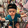 baby, shopping_cart, hat, clothing_store, baby_clothes, blue_onesie, person, child, retail, backwards_cap, shopping, cute, wide_eyes, indoor, rack, apparel, infant, store, toddler, curious