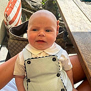 Georgi is registered to the contest to win money with this photo: baby, infant, child, outdoor, person, clothing, white_shirt, overalls, buttons, face, head, lap, pillow, striped_pillow, table, wooden_table, greenery, sunlight, hand, sitting
