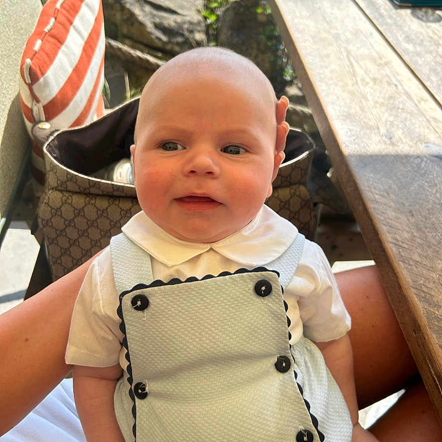 Georgi is registered to the contest to win money with this photo: baby, buttons, child, clothing, face, greenery, hand, head, infant, lap, outdoor, overalls, person, pillow, sitting, striped_pillow, sunlight, table, white_shirt, wooden_table