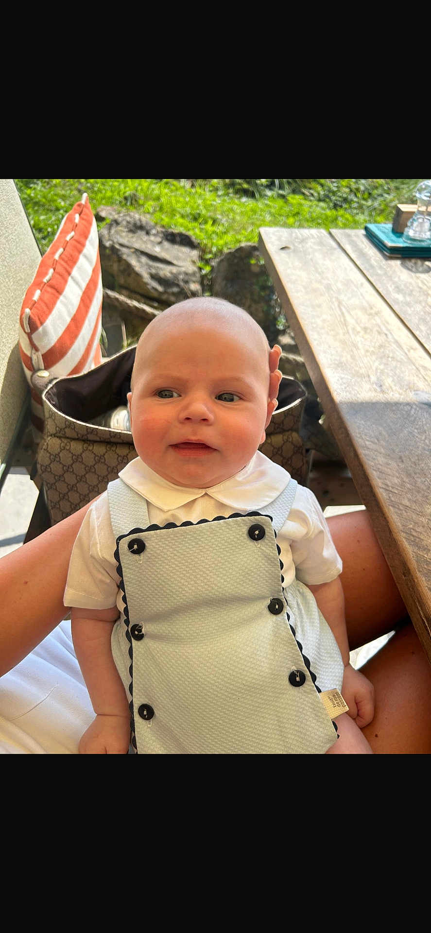Georgi is registered to the contest to win money with this photo: baby, infant, child, outdoor, person, clothing, white_shirt, overalls, buttons, face, head, lap, pillow, striped_pillow, table, wooden_table, greenery, sunlight, hand, sitting