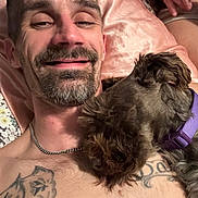 Hazel is registered to the contest to win money with this photo: man, dog, pet, sleeping, collar, tattoo, chain_necklace, bed, pillow, closeup, facial_hair, smile, skin, resting, indoors, human, animal, brown_dog, purple_collar, comfort