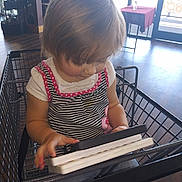 Gina is registered to the contest to win money with this photo: child, toddler, baby, sitting, shopping_cart, indoor, store, supermarket, tablet, book, reading, blonde_hair, hair, face, clothing, striped_shirt, polka_dots, portrait, wooden_floor, window