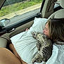 cat, kitten, woman, person, pillow, car_interior, passenger_seat, seatbelt, window, side_mirror, road, guardrail, greenery, sleeping, nap, cozy, striped_fur, hand, t_shirt, travel