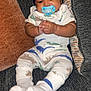 baby, infant, pacifier, hat, dinosaur_print, onesie, socks, couch, pillow, clothing, hands, face, smiling, seated, indoor, cute, child, young, cozy, comfort