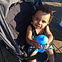 toddler, child, stroller, sippy_cup, outdoor, sunlight, wooden_deck, black_tank_top, blue_shorts, sandals, curly_hair, smiling, baby, seatbelt, shadow, person, playful, summer, daylight, cute