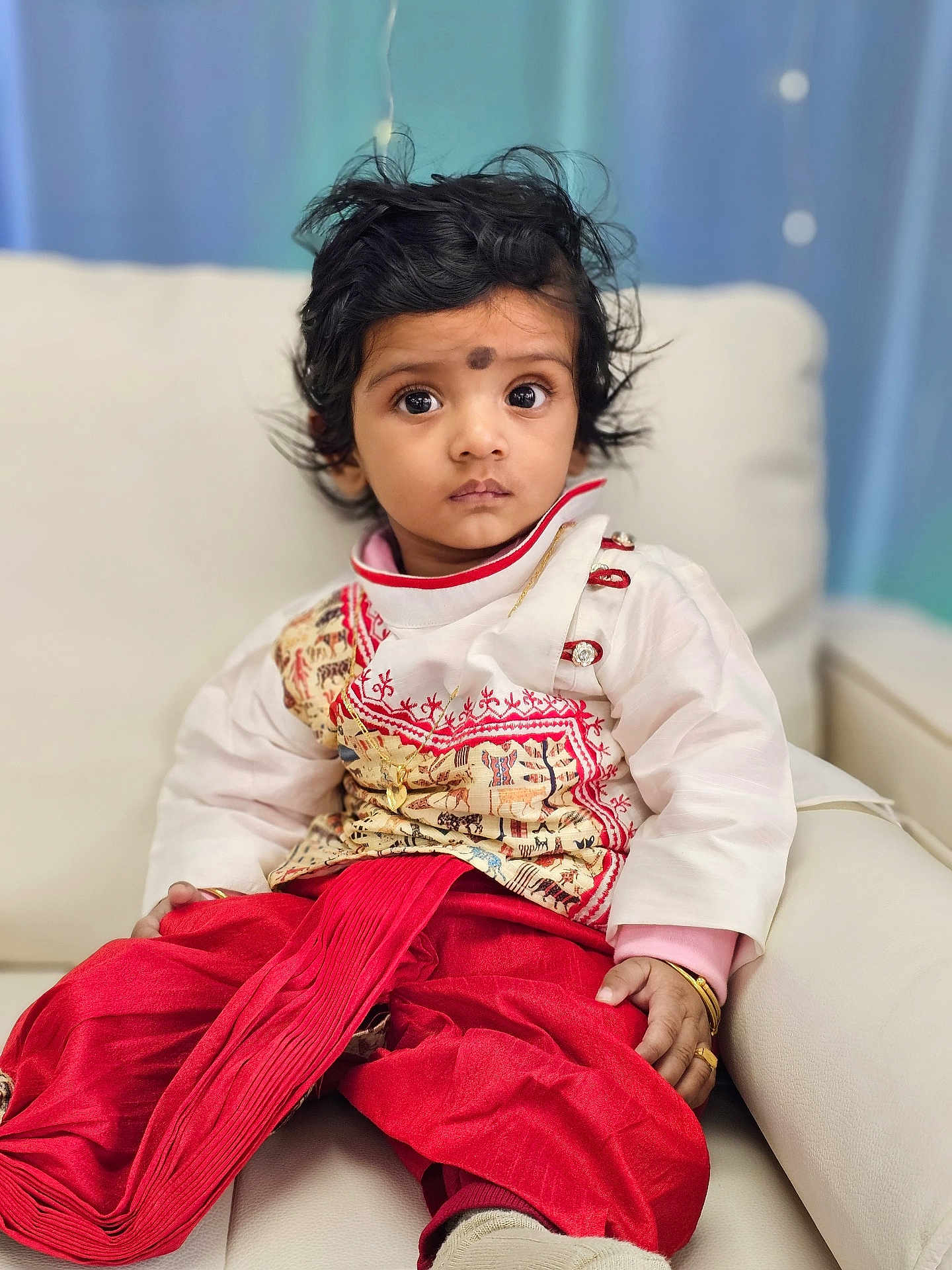 Sanvik is registered to the contest to win money with this photo: toddler, child, portrait, indoors, traditional_clothing, red_pants, white_top, jewelry, bracelet, ring, couch, sofa, curtain, face, hair, expression, sitting, young_child, cute, person
