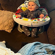 Grayson is registered to the contest to win money with this photo: baby, child, infant, cap, backwards_cap, activity_seat, toy, blanket, couch, brown_couch, smile, happy, indoor, cozy, plush, clothing, footed_pajamas, person, seated, face