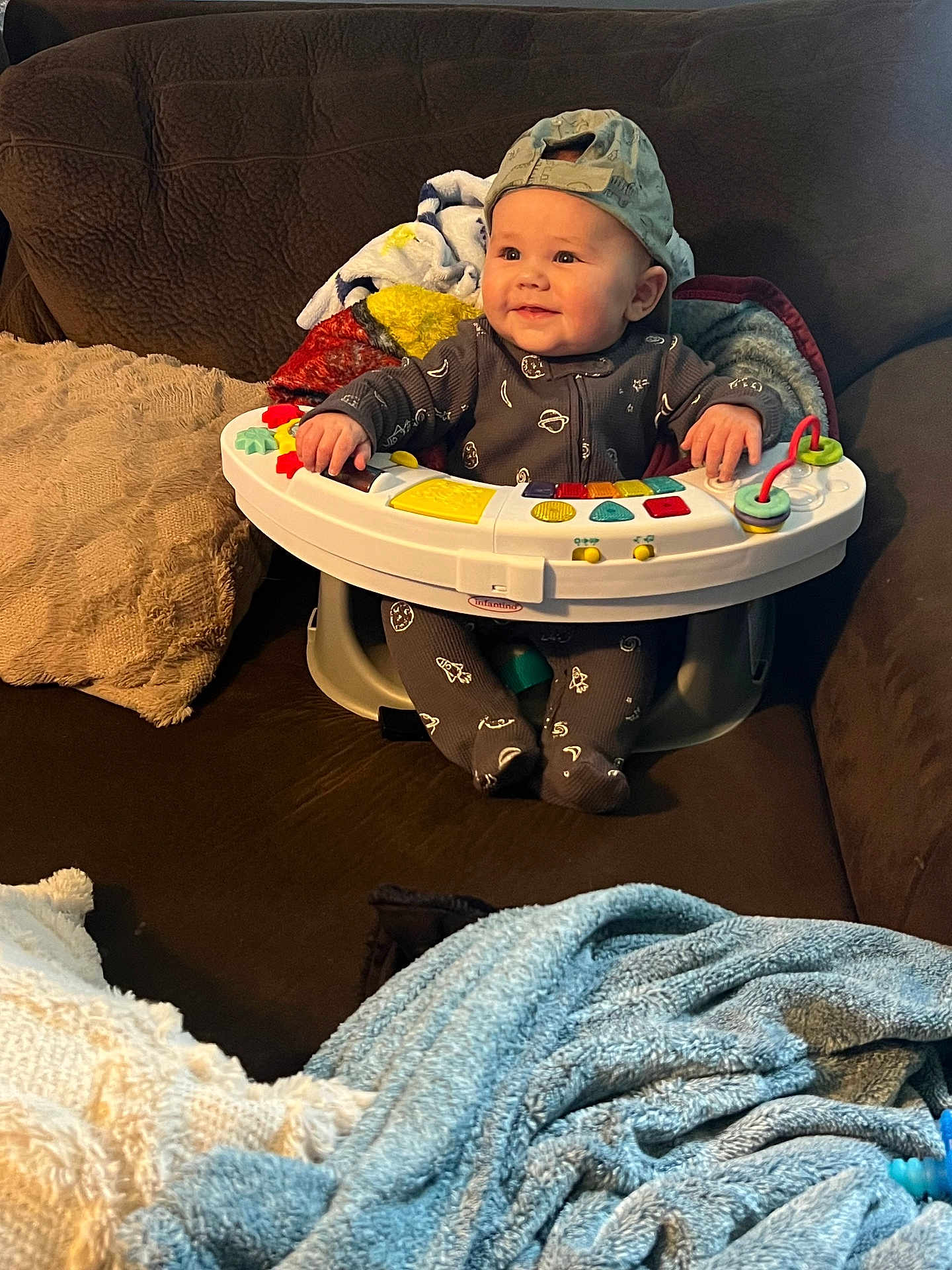 Grayson is registered to the contest to win money with this photo: baby, child, infant, cap, backwards_cap, activity_seat, toy, blanket, couch, brown_couch, smile, happy, indoor, cozy, plush, clothing, footed_pajamas, person, seated, face