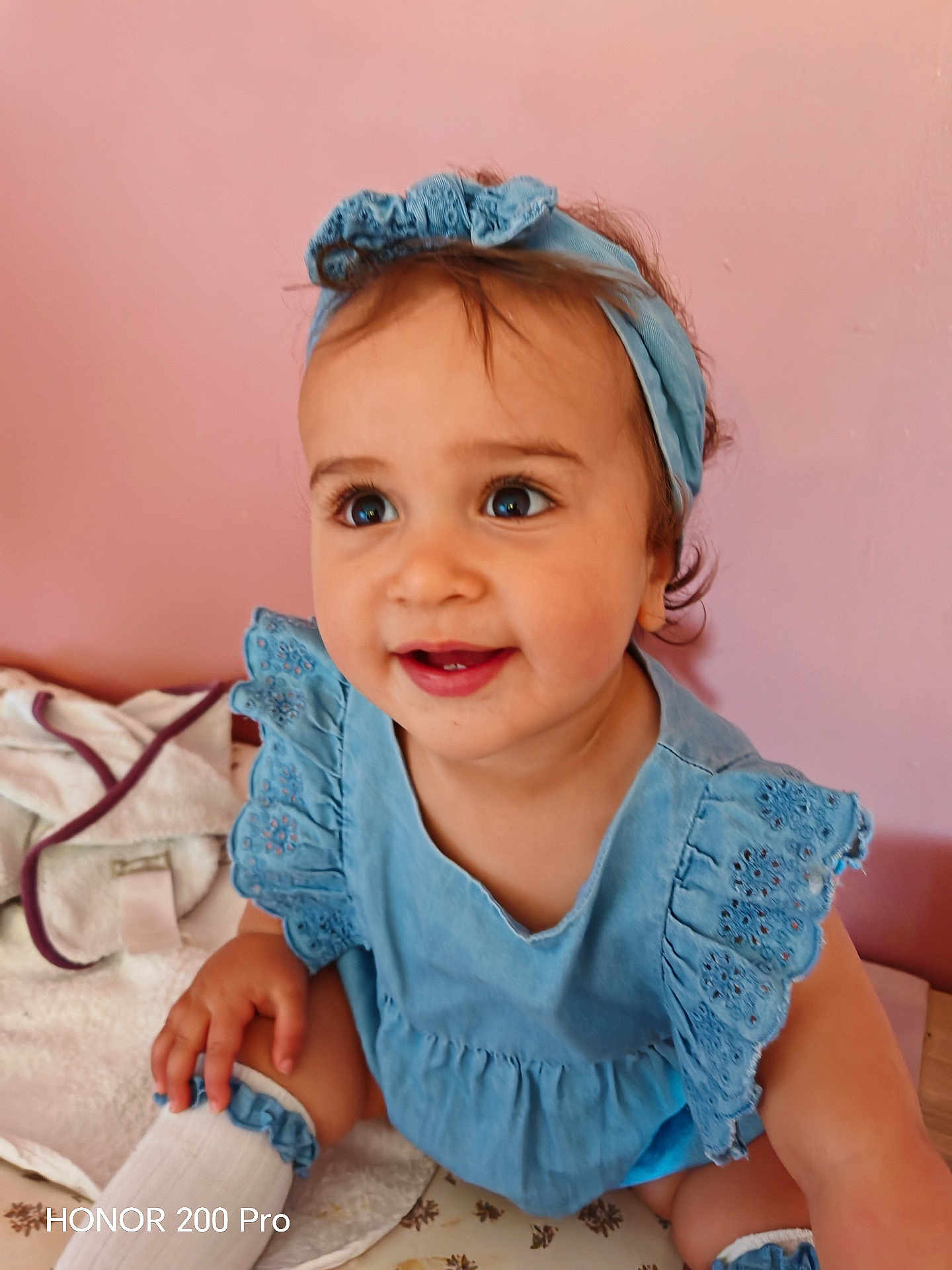 Lya participe au concours pour gagner de l'argent avec cette photo : baby, child, blue_clothing, headband, smile, happy, indoor, pink_wall, cute, sitting, infant, person, portrait, young_child, dressed_up, hand, face, eyes, skin, expression